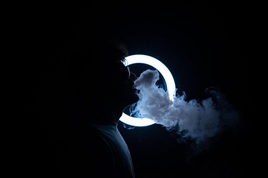 Silhouette of a man exhaling smoke illuminated by a ring light in a dark room.