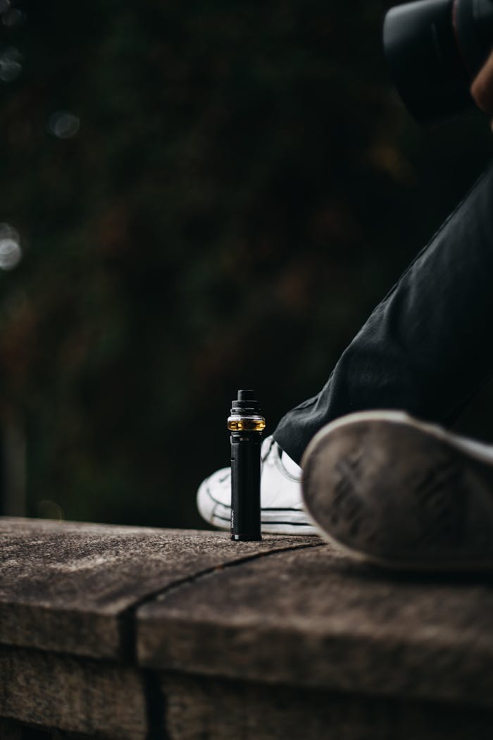 A person wearing sneakers sits outdoors with a vape device on a wooden ledge.
